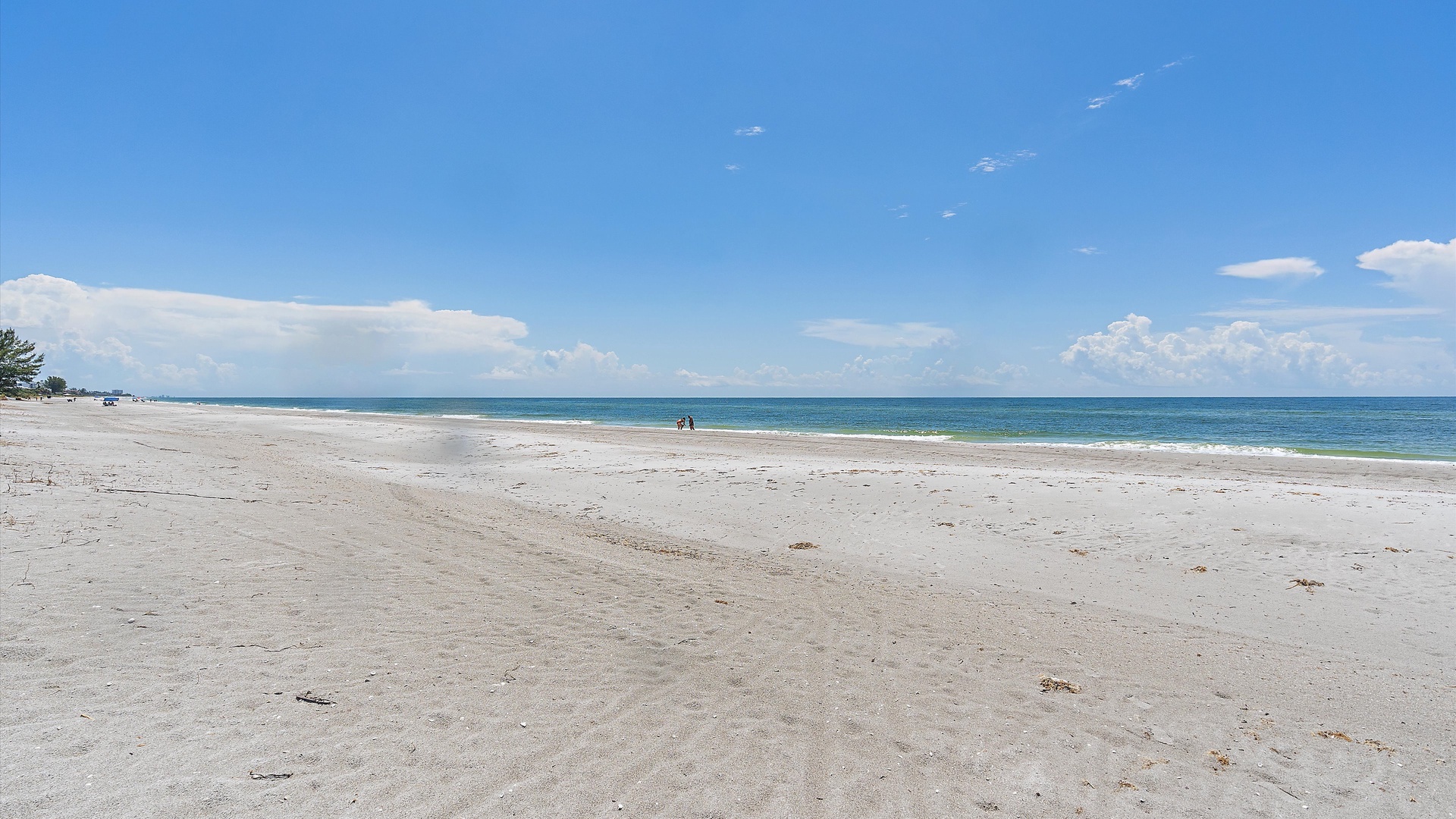 A vast, empty sandy beach stretches into the distance under a clear blue sky, with the calm sea meeting the horizon.