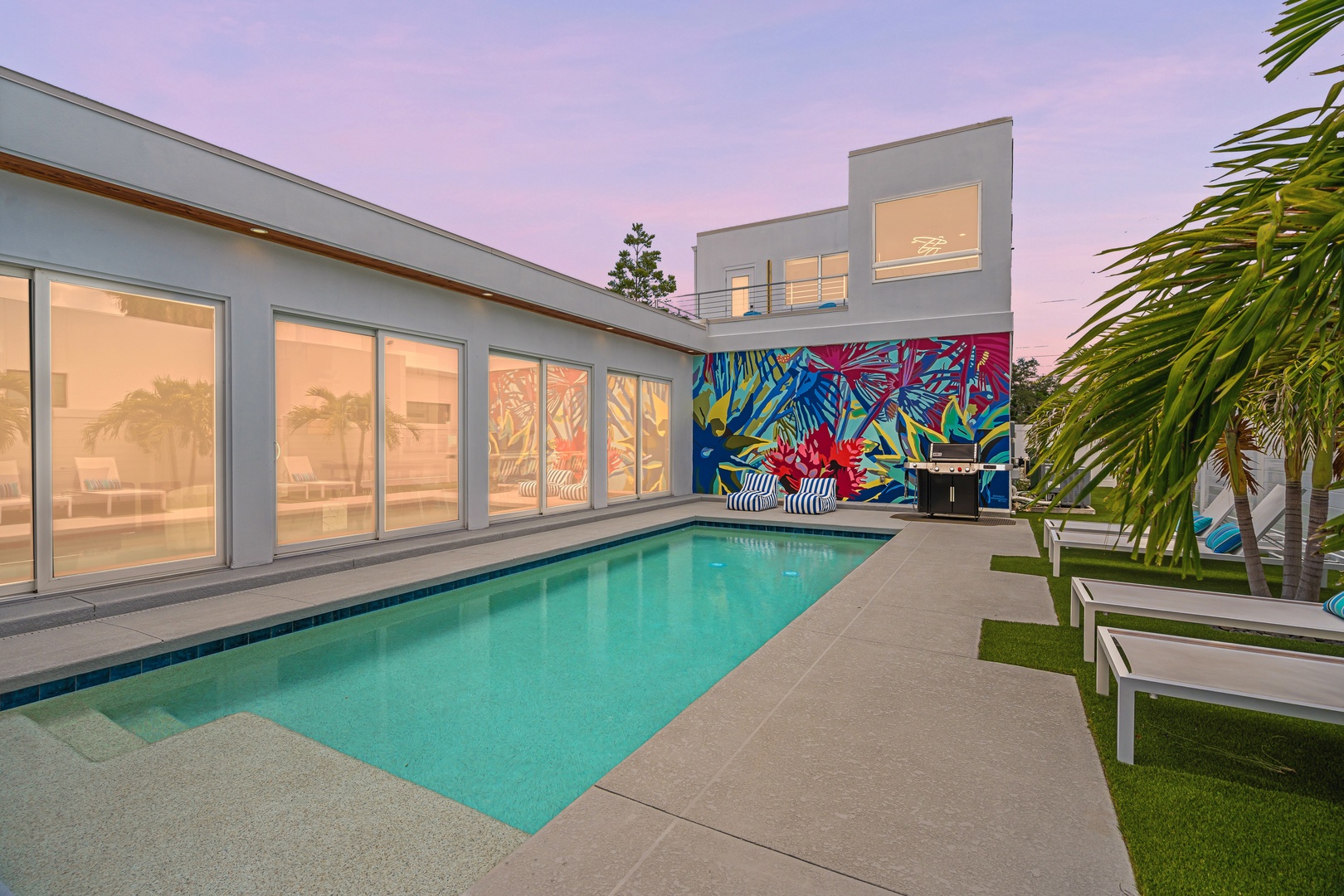 Modern vacation retreat featuring a sparkling pool, vibrant tropical murals, and sleek architecture, framed by palm trees under a stunning pink dusk sky.