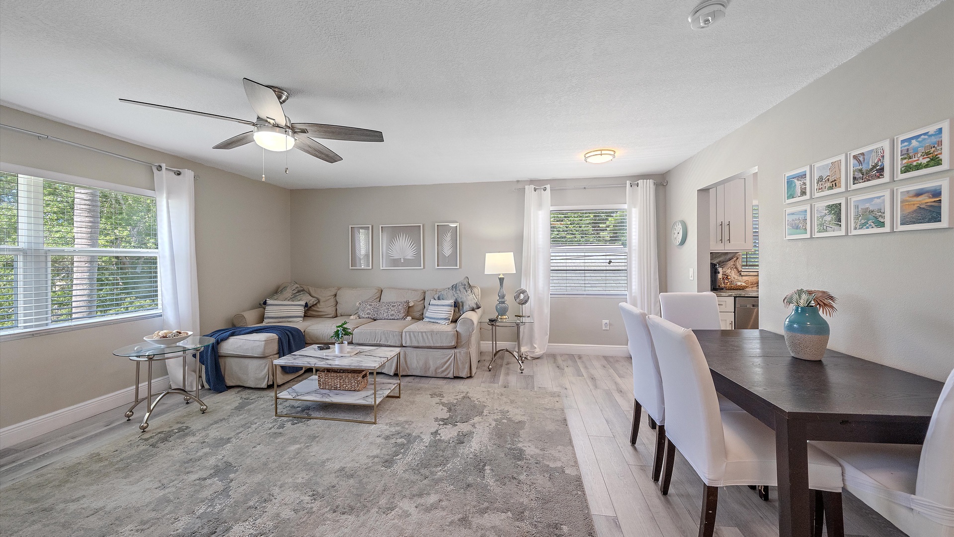 Relax in your open-concept living and dining space with serene coastal touches, plush seating, and plenty of natural light streaming through large windows overlooking lush greenery.