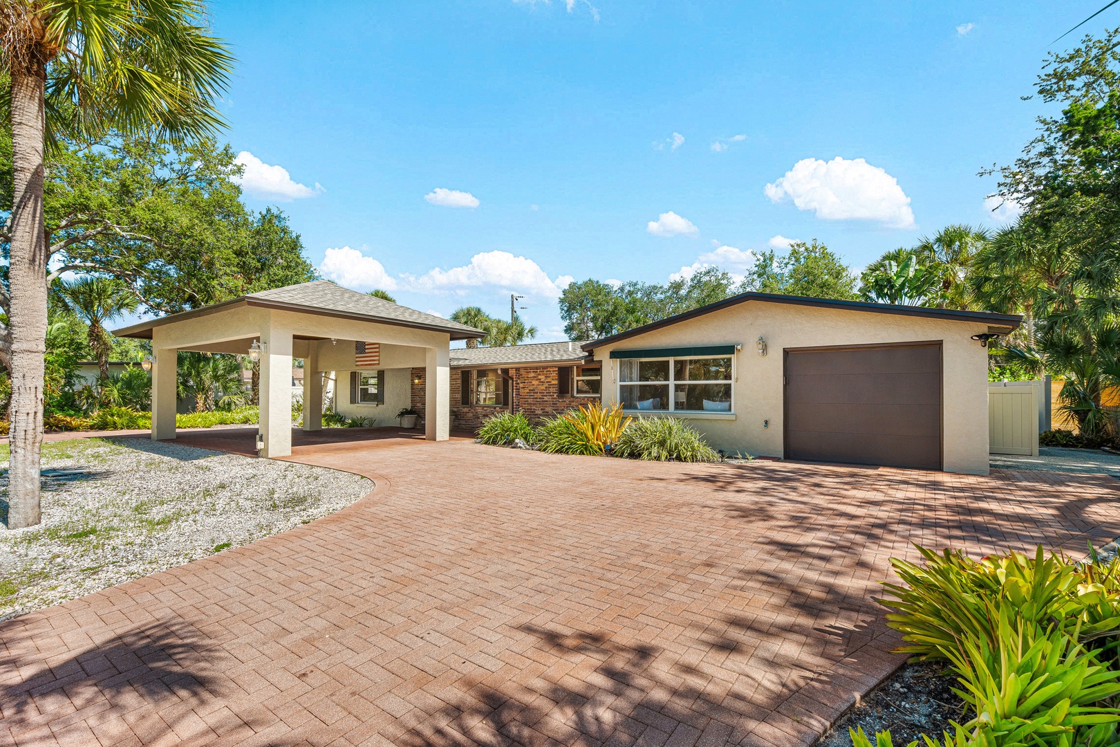 A beautifully landscaped vacation home features a covered carport and attached garage, surrounded by palm trees and tropical greenery under bright Florida skies.