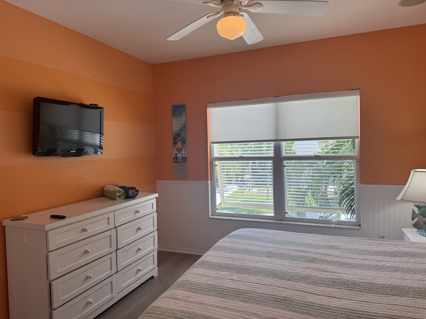 Your serene bedroom features warm coral tones, a ceiling fan to keep you comfortable, and natural light streaming through the window. Relax while enjoying your favorite shows—it's your perfect tropical retreat.