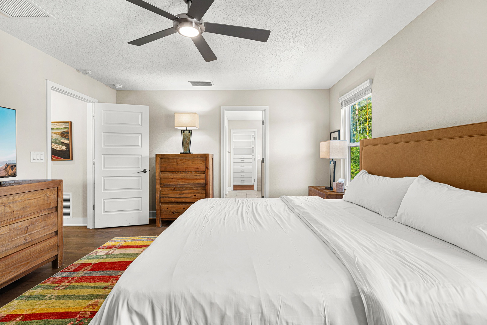 Drift into peaceful slumber beneath the gentle breeze of a ceiling fan, wrapped in plush white linens. Warm wood furnishings and a vibrant rug add character, while natural light from the window brightens your mornings.