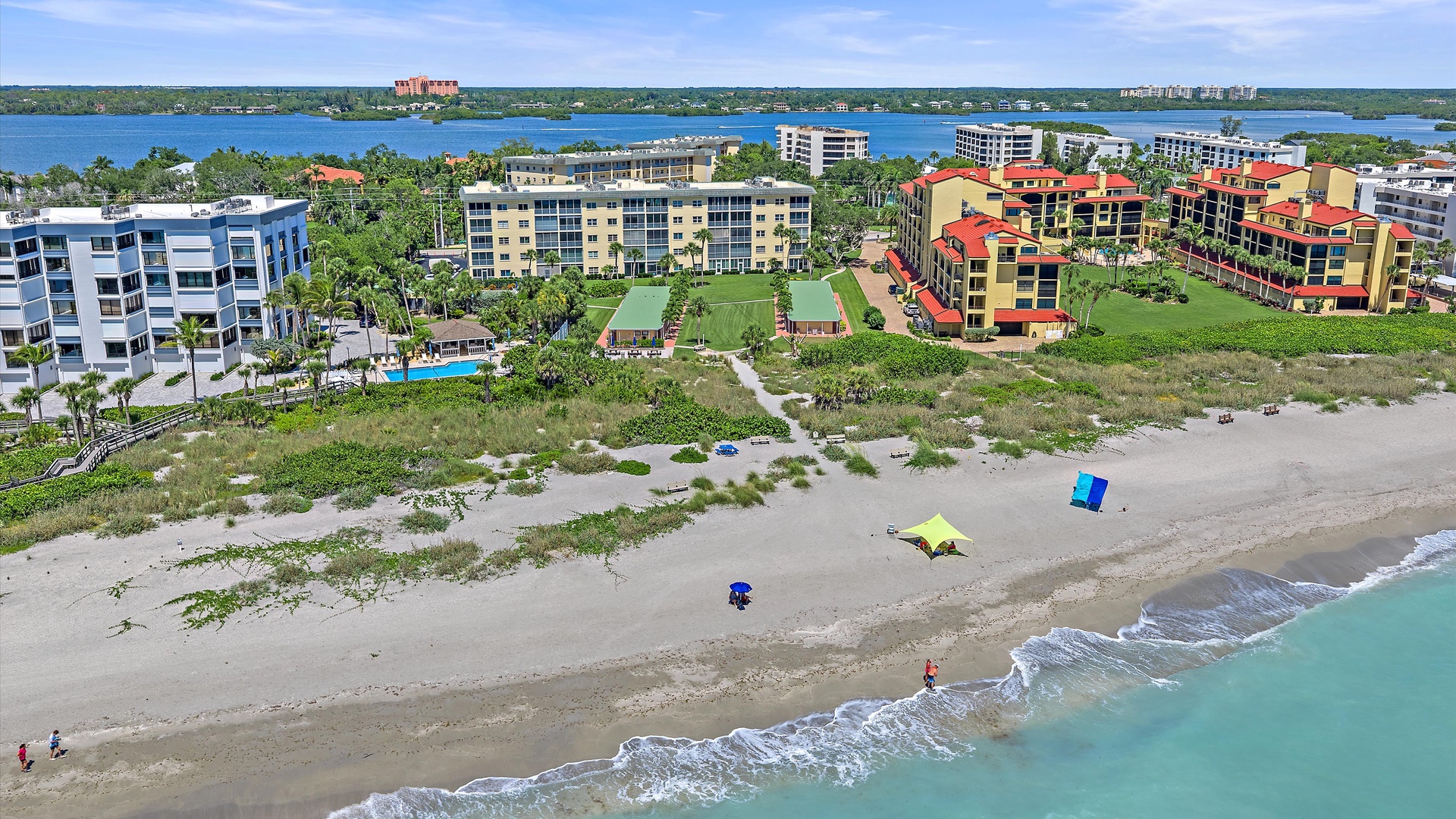 Aerial view of beachfront property nestled between pristine sandy shores and calm bay waters, featuring colorful low-rise buildings surrounded by lush tropical landscaping and waterfront access.