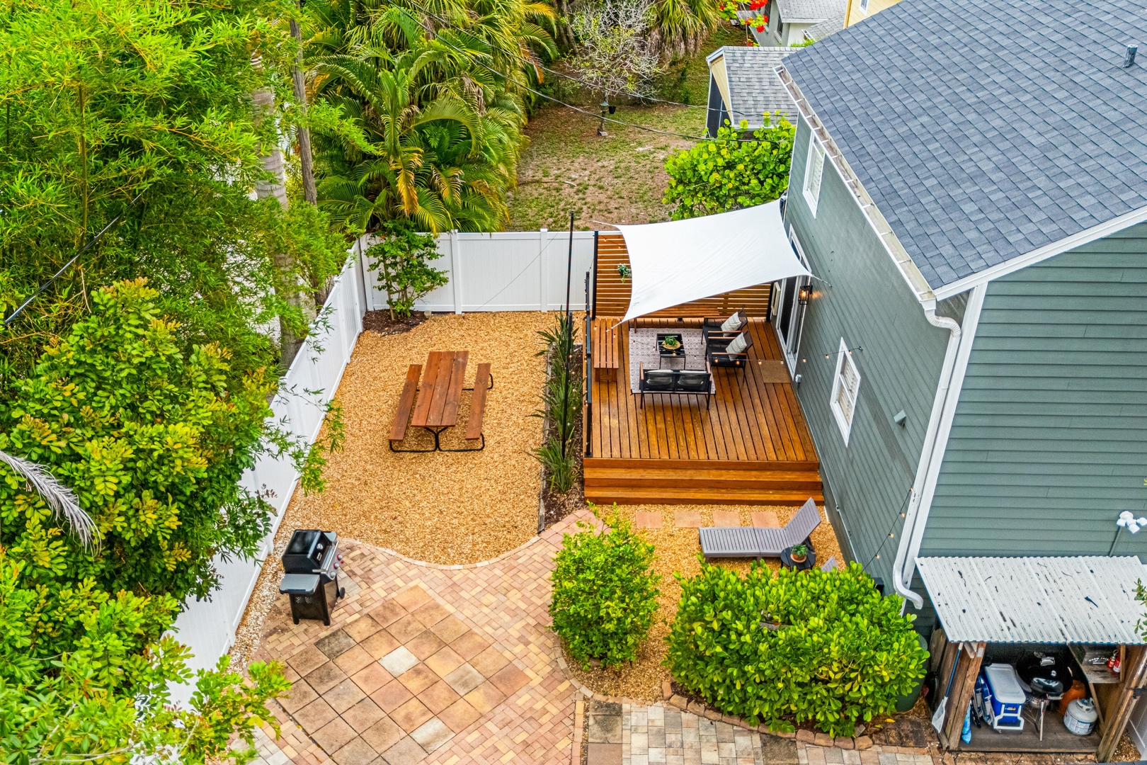 Aerial view showcasing the property's backyard with wooden deck, shaded seating area, picnic table, BBQ grill, and lush surrounding greenery.
