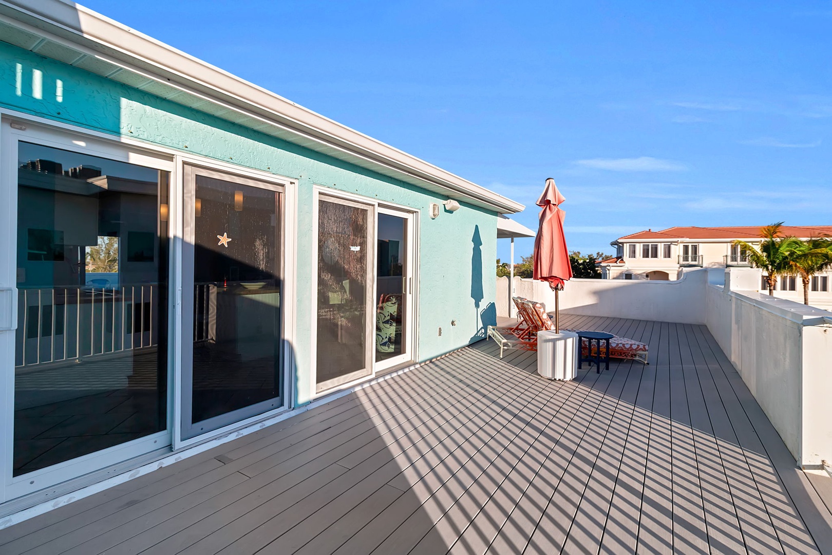The spacious rooftop deck features coastal-inspired turquoise exterior walls and offers generous outdoor living space with a sun umbrella and seating, perfect for relaxing under clear blue skies in a peaceful residential setting.