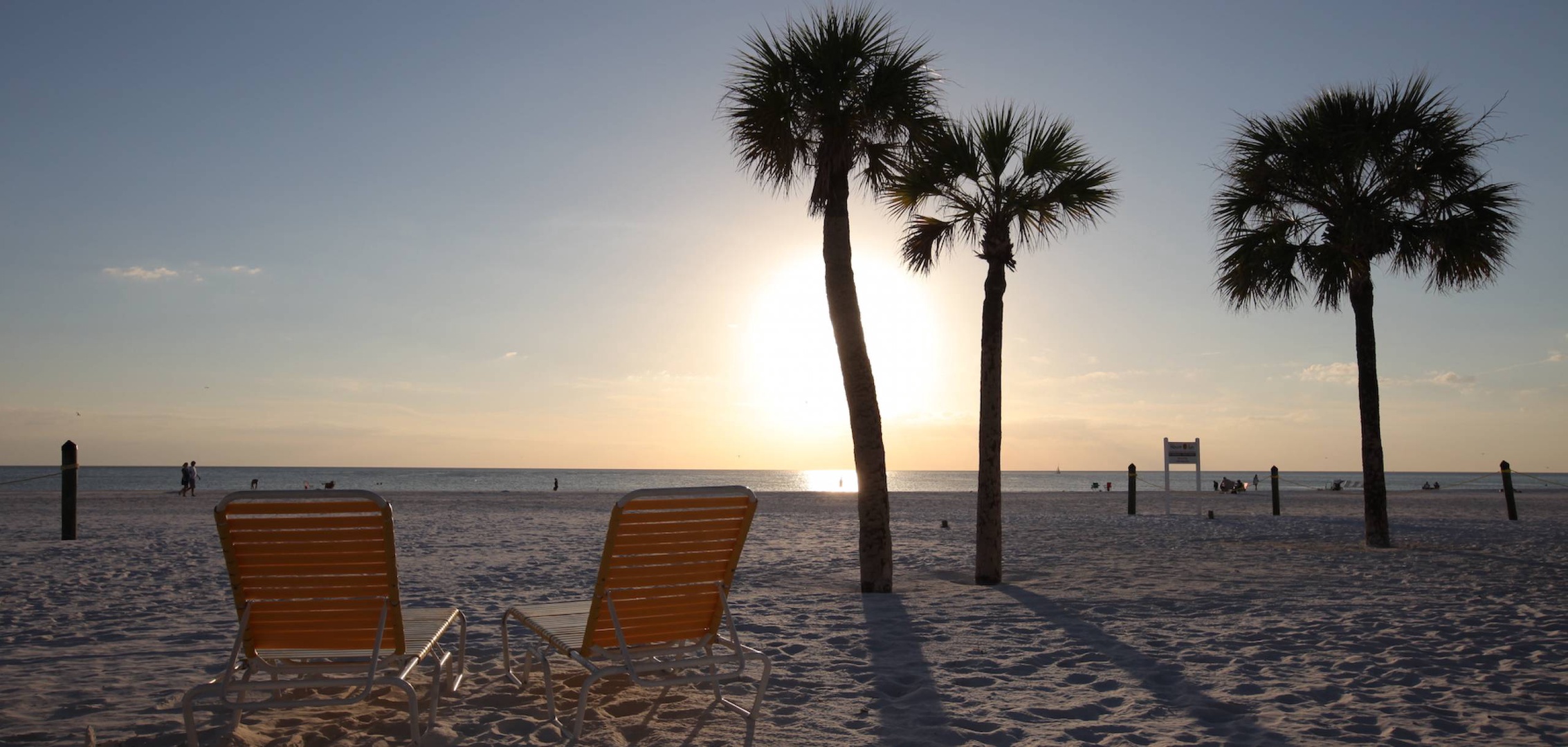 Two beach chairs face the Gulf of Mexico beneath swaying palm trees as the sun sets over the water, creating the perfect scene for your evening relaxation.