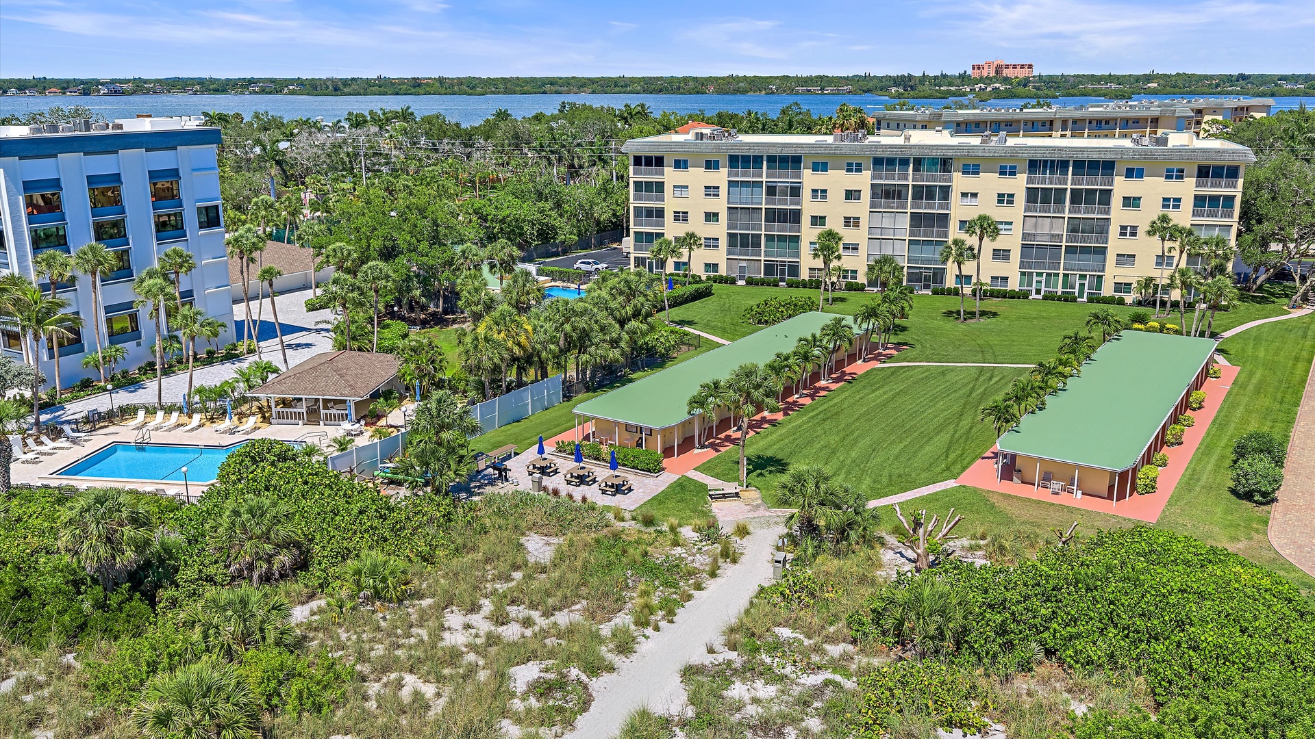 An aerial view showcasing the property complex surrounded by lush tropical landscaping, waterfront proximity, and neighboring residential buildings in a scenic coastal setting.