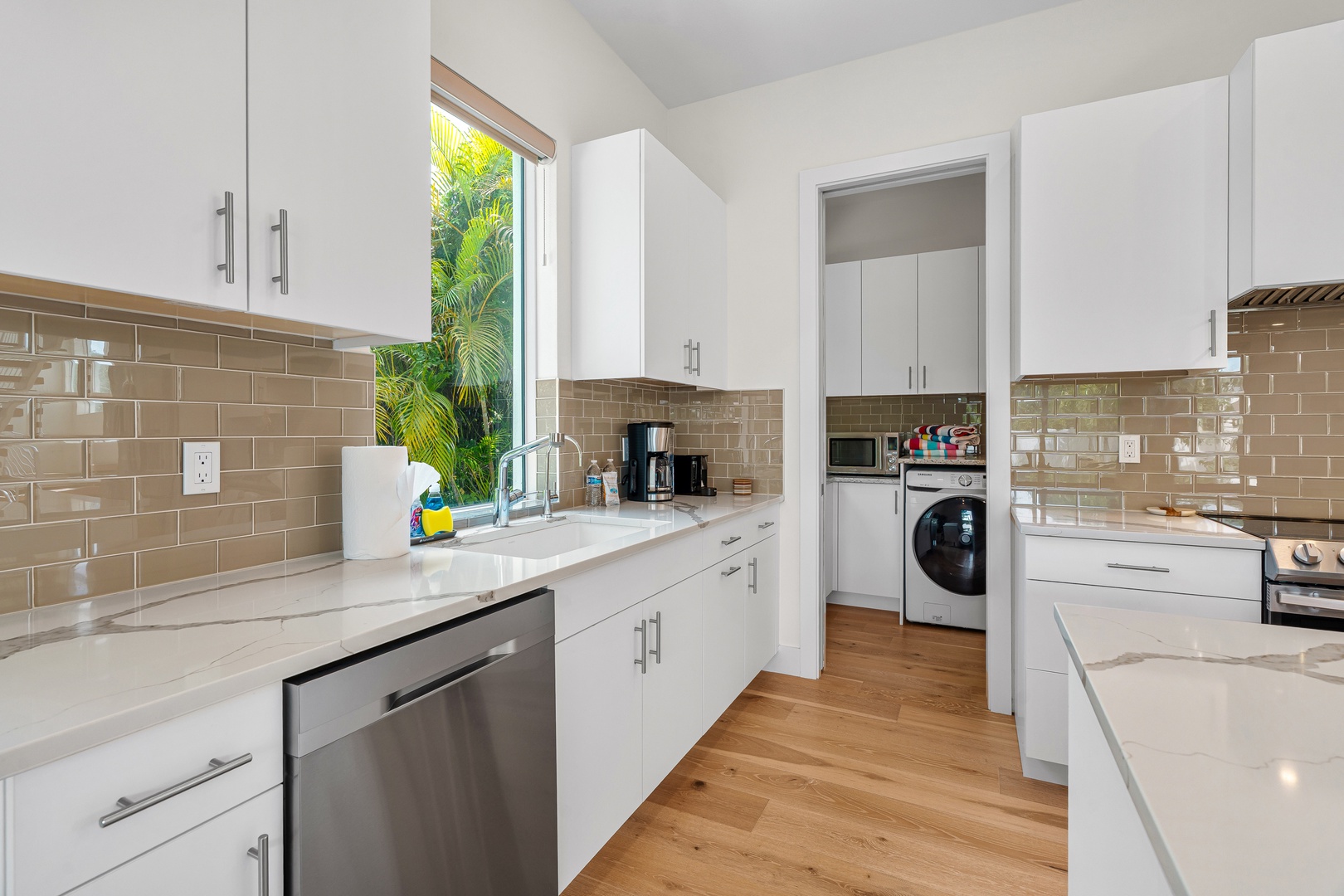 Your fully equipped kitchen with sleek white cabinetry, glossy tile backsplash, and modern appliances makes meal prep a breeze while lush greenery outside the window adds a tropical touch.