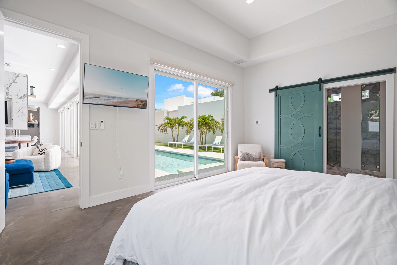 Step into your serene bedroom retreat, where crisp white linens and a charming barn door invite relaxation. Sliding glass doors reveal a sparkling pool and tropical palms just steps away—your perfect escape.
