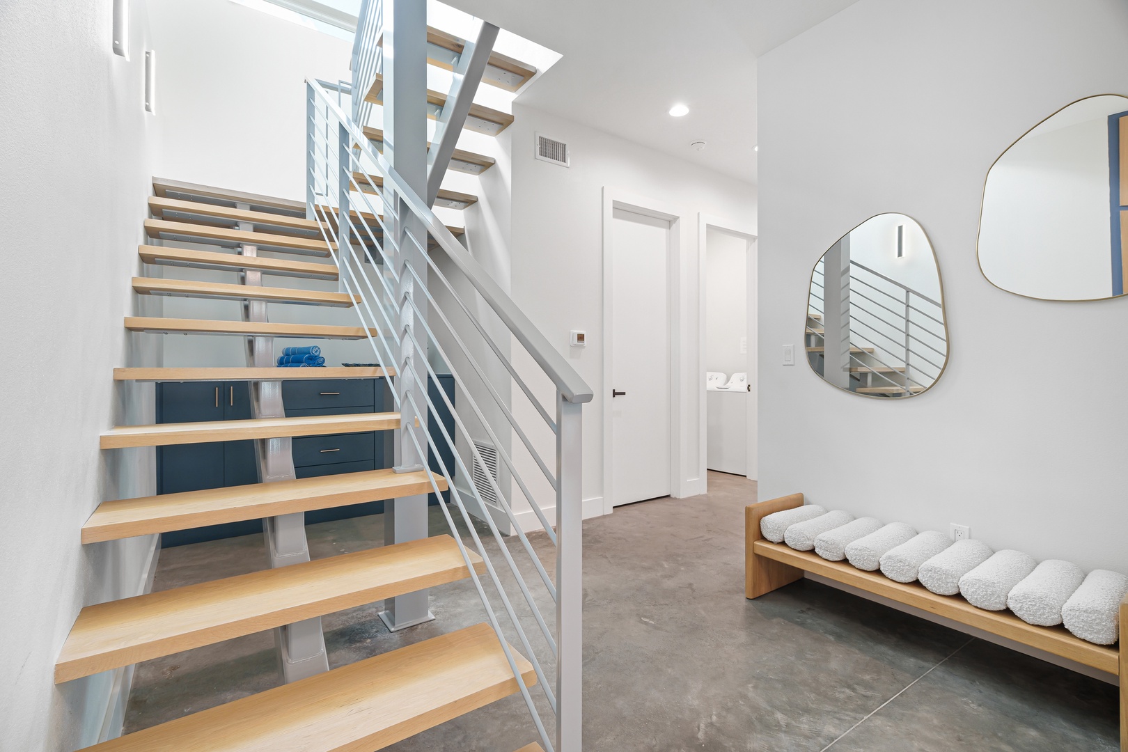 Open staircase with natural wood steps and sleek metal railings leads to your upper-level spaces, while modern mirrors and a cushioned bench create a welcoming entry area.