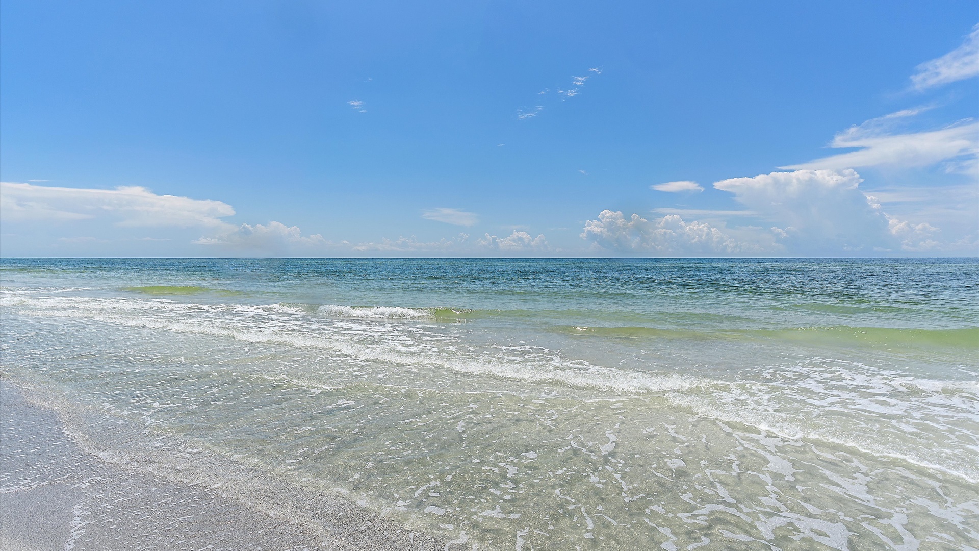 A tranquil beach scene with gentle waves lapping onto the shore and a clear blue sky with scattered clouds above.