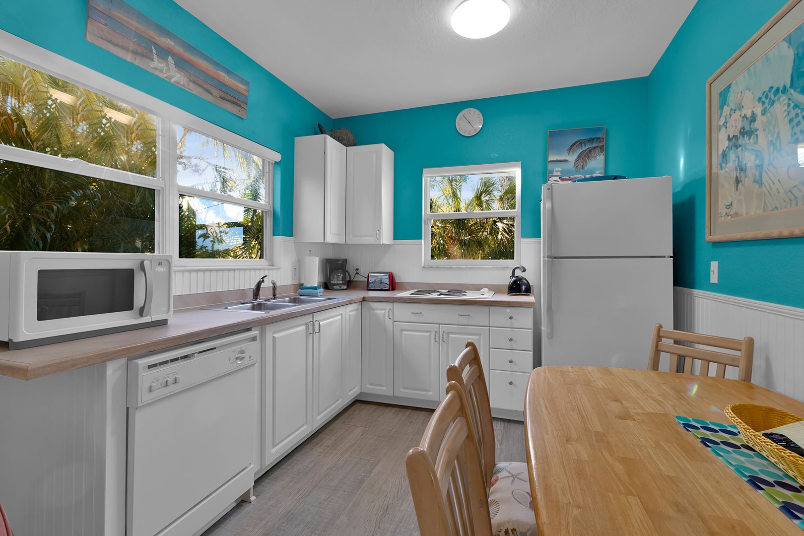 Your fully equipped kitchen features bright turquoise walls and natural light streaming through palm-framed windows, creating a cheerful coastal vibe for preparing meals during your stay.