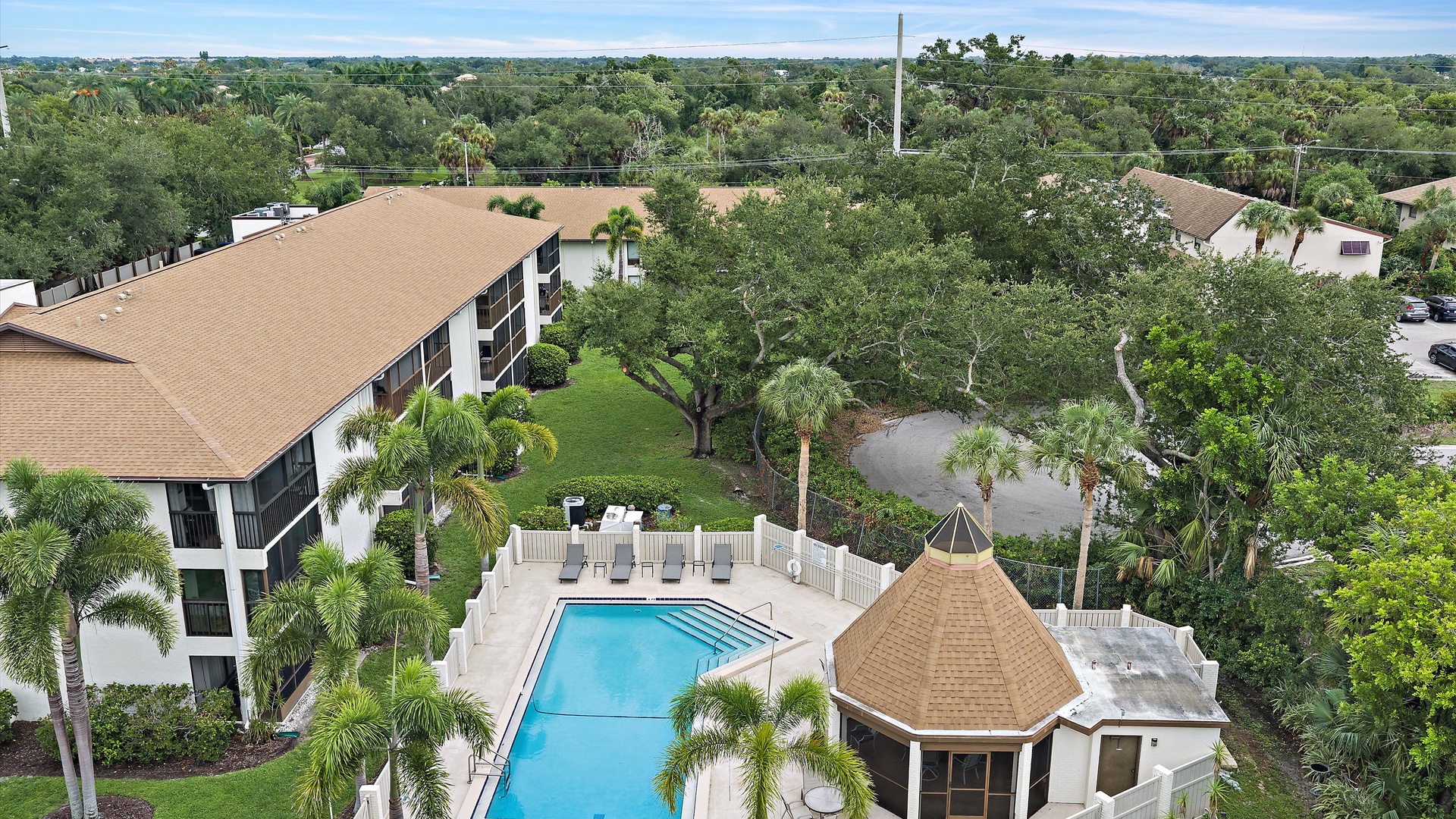 The property sits nestled in a lush, tropical residential setting with palm trees and mature greenery visible throughout the surrounding area. A welcoming community pool and gathering spaces are centrally located within the landscaped grounds.