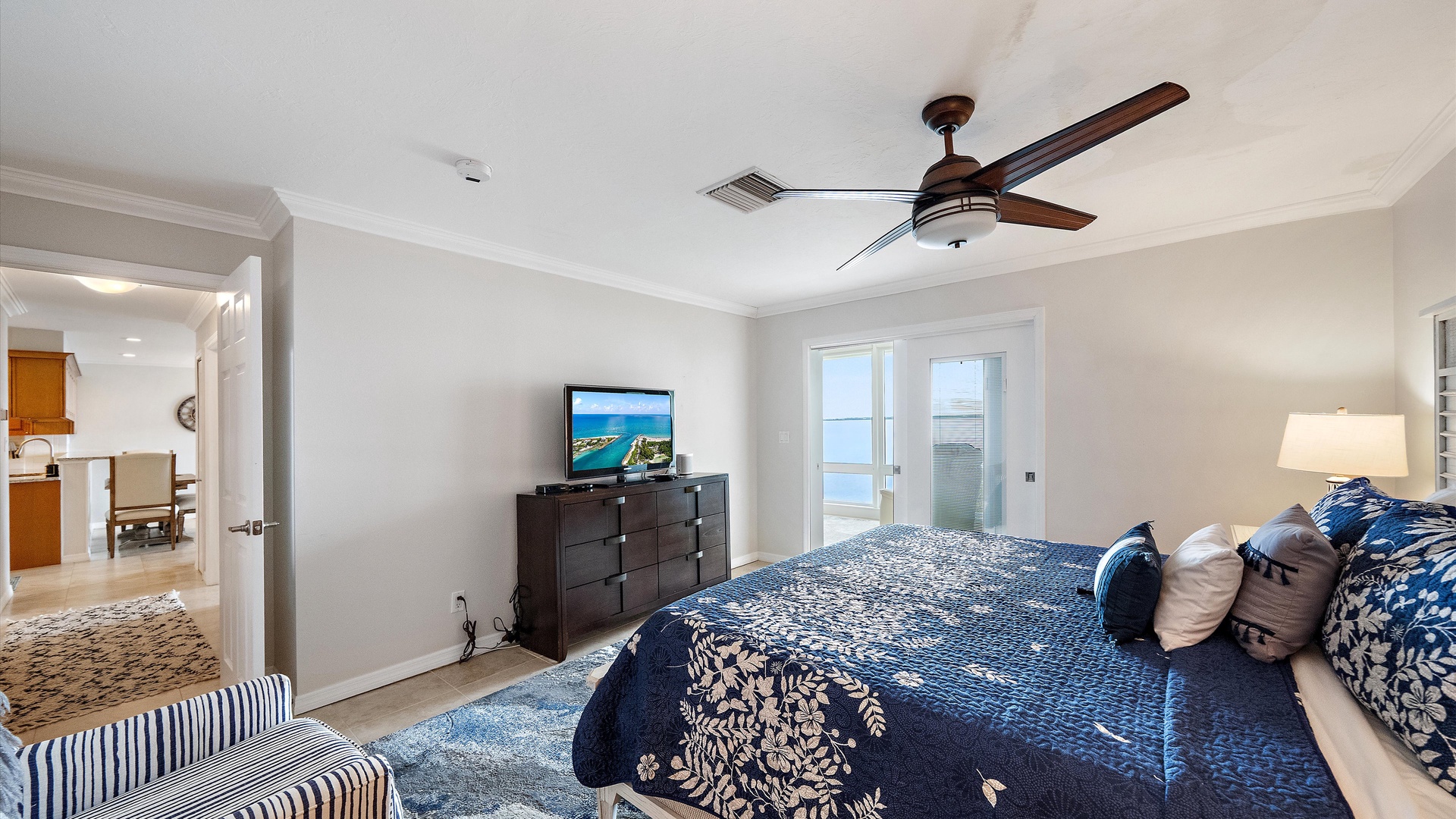 The primary bedroom pairs a king bed with its own television and access to the enclosed sunroom. Blue-and-white bedding and coastal furnishings create a comfortable retreat beside the bay.