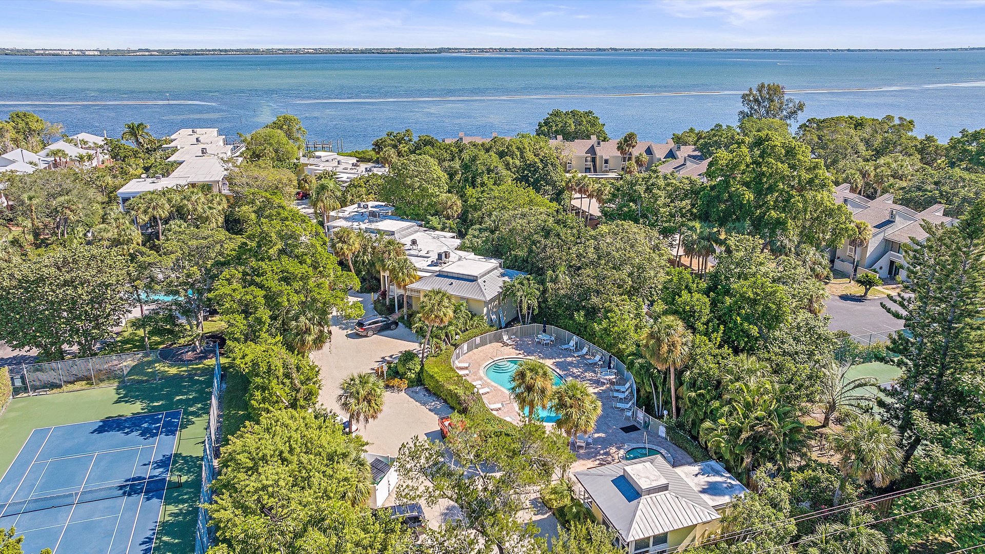 An aerial view of a coastal resort featuring a swimming pool, tennis court, and surrounding greenery, with a large body of water in the background.