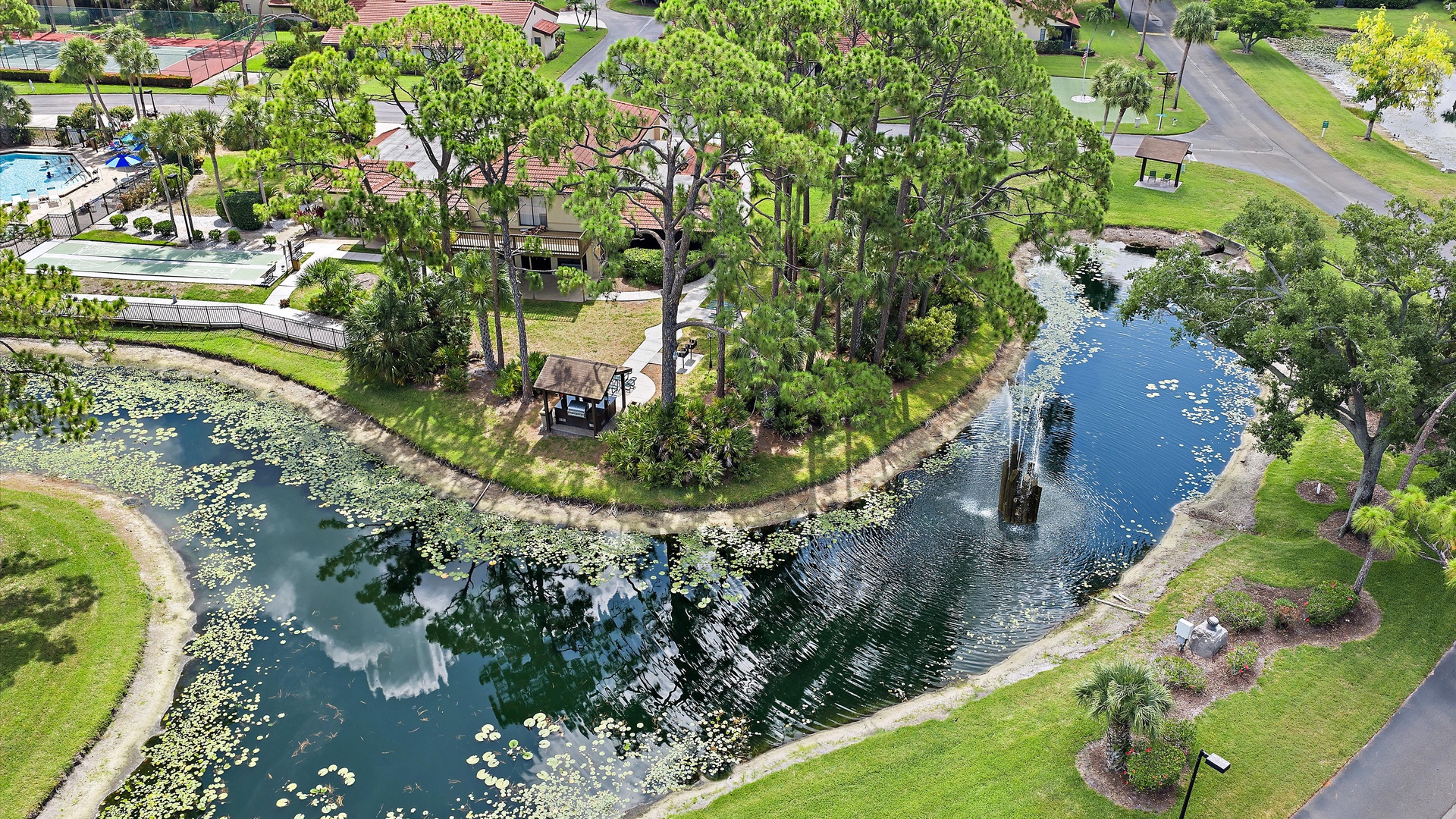 Aerial view of the property nestled within a peaceful residential community, featuring a tranquil pond, mature trees, and well-maintained grounds with swimming pool amenities nearby.