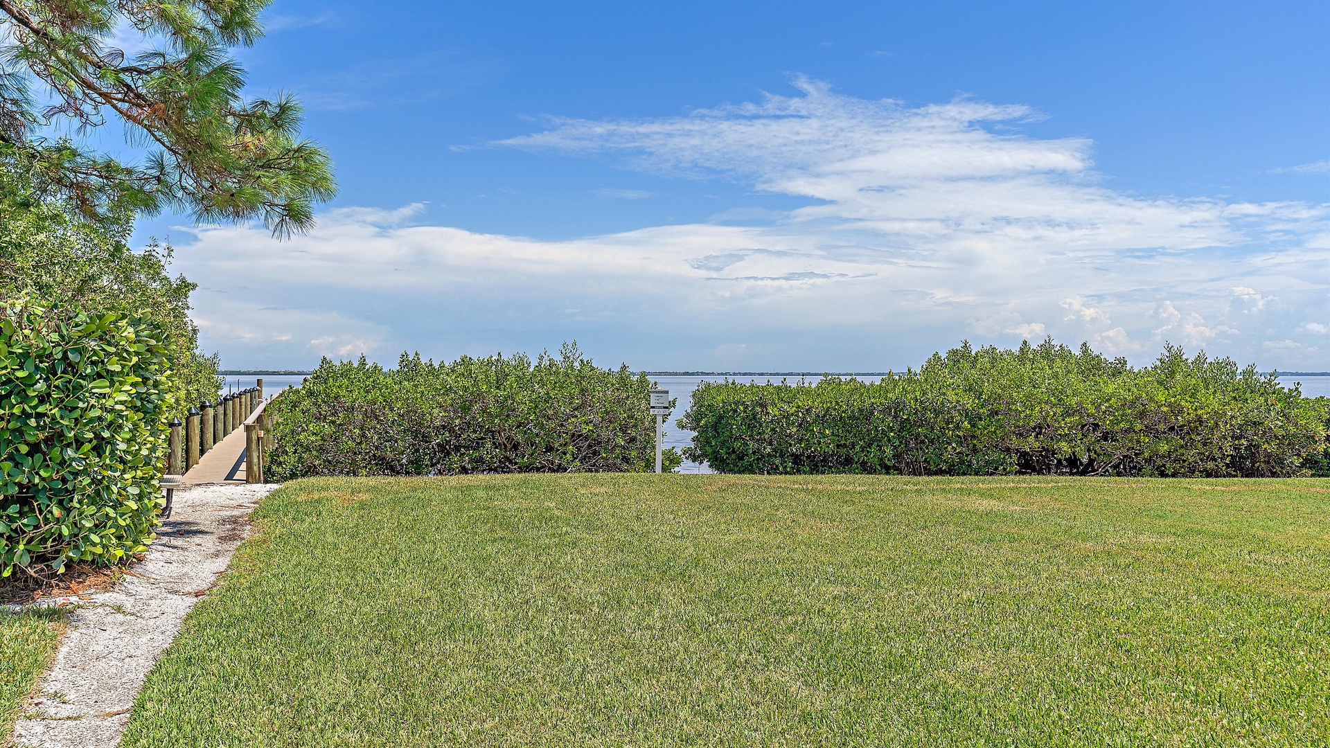 A grassy yard with a path leading to a dock surrounded by lush greenery, overlooking a calm body of water and a blue, partly cloudy sky.