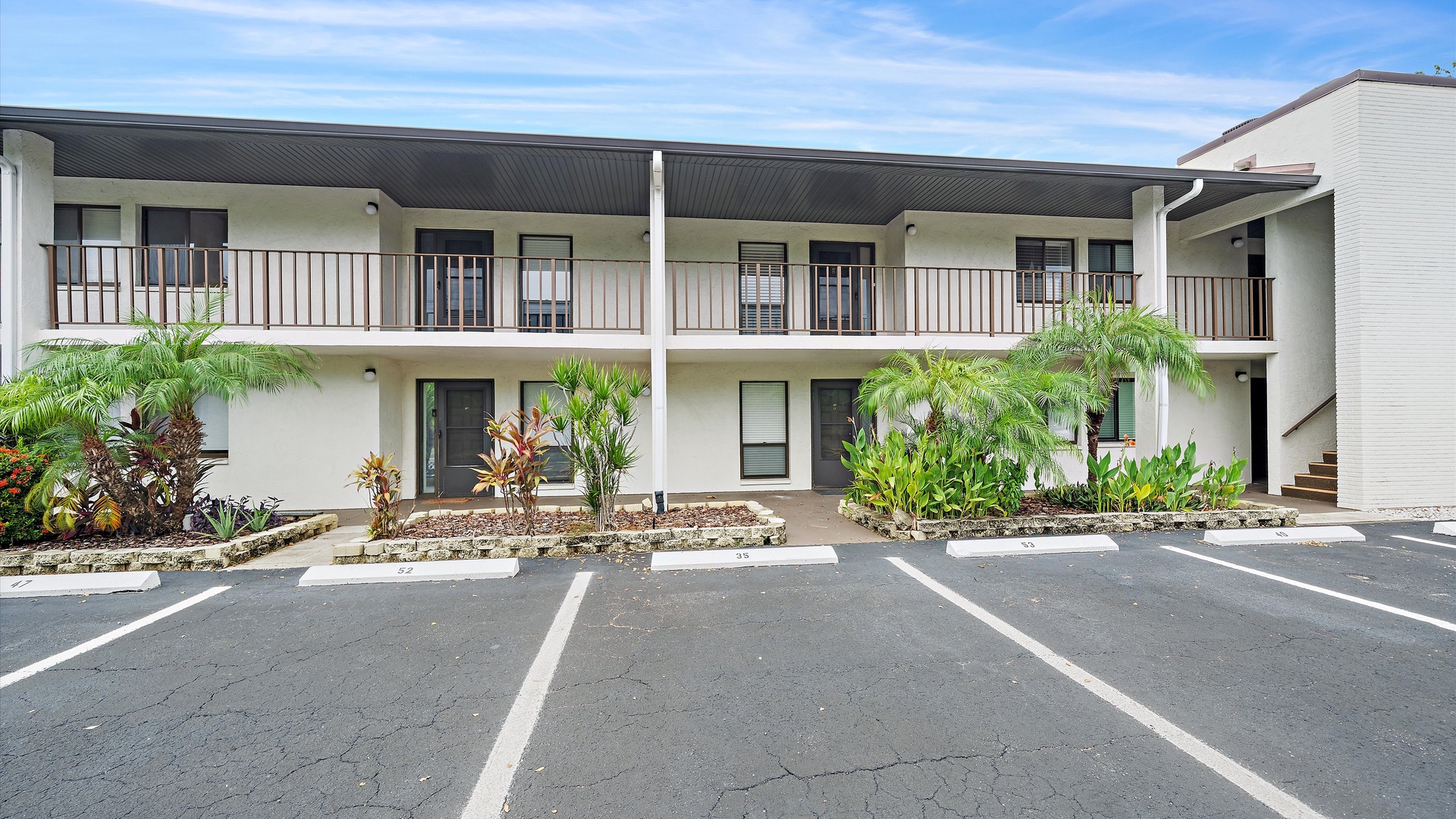 A two-story building with private balconies framed by lush tropical palms, set within a well-maintained property featuring convenient parking spaces and landscaped island beds.