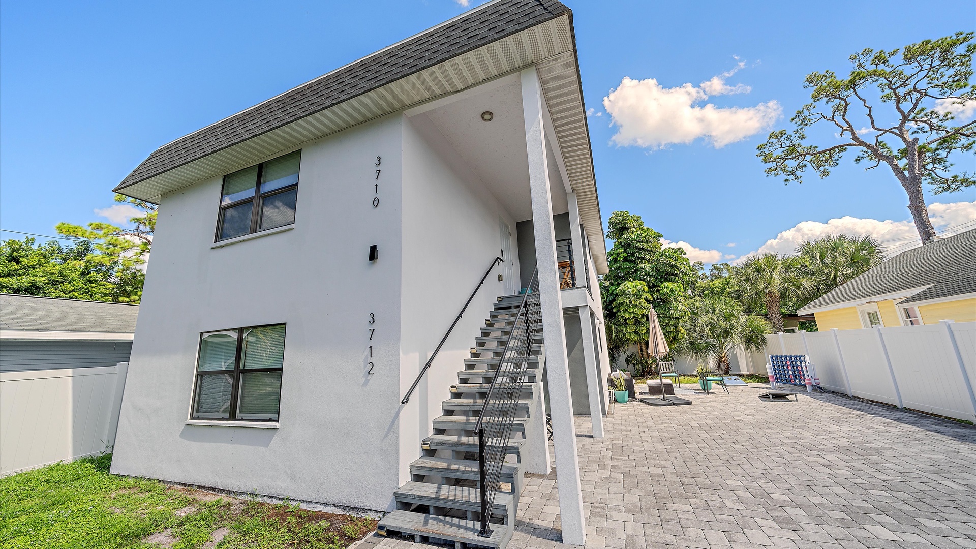 Modern two-story building with a spacious courtyard, featuring open staircases and a paver patio. Palm trees and tropical greenery frame the property in this sunny coastal setting.