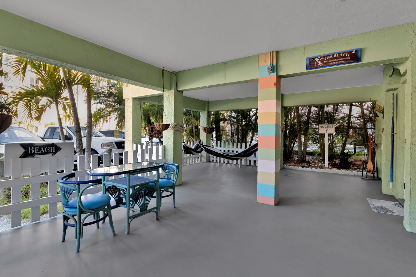 A covered outdoor area leads to the beach, with colorful striped pillars, hammocks strung for relaxation, and palm trees swaying beyond the white picket fence.