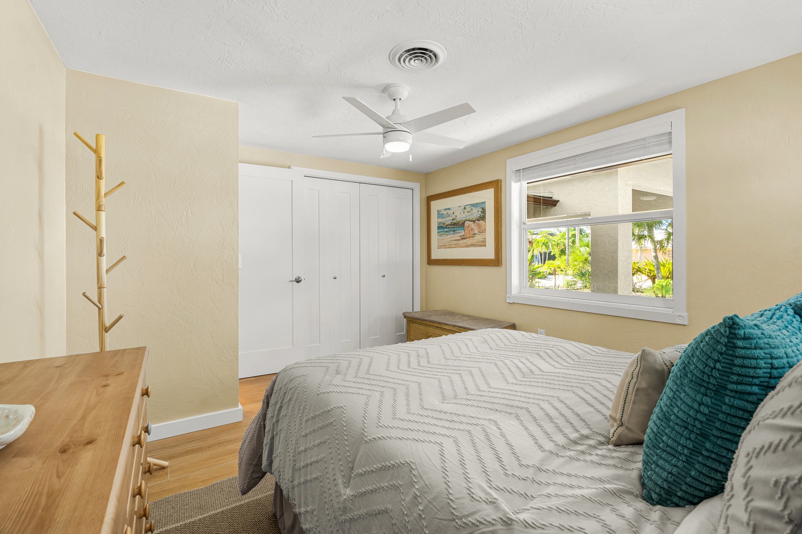 Relax in this peaceful bedroom with a cozy bed, ceiling fan, and natural light streaming through the window overlooking tropical greenery. Your coastal retreat awaits.