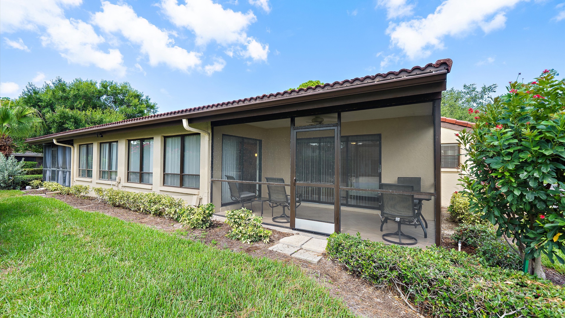 Well-maintained property nestled among lush landscaping and tropical greenery. Relax on the screened patio with comfortable seating, surrounded by gardens under blue skies.