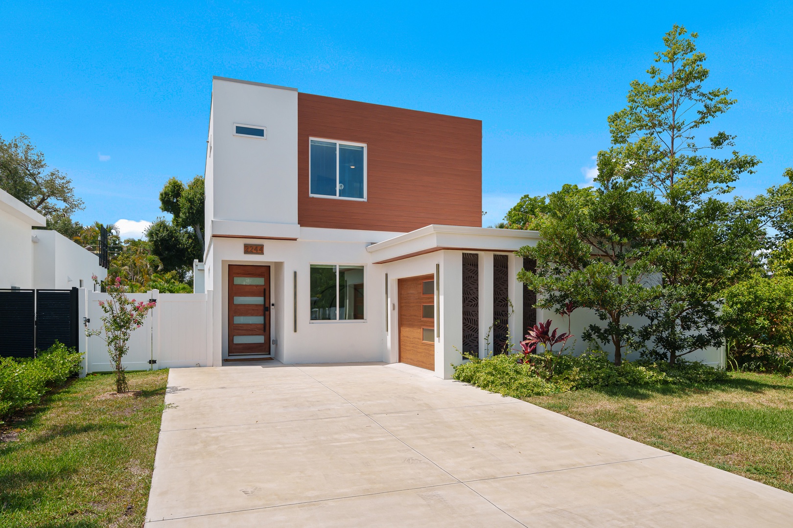 Modern two-story vacation rental featuring contemporary architecture with wood accents, set in a peaceful residential area surrounded by lush tropical trees and greenery under clear blue skies.