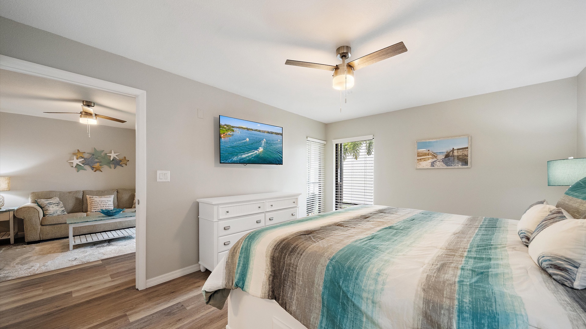 Relax in your bright, coastal-inspired guest bedroom with a plush queen bed, ceiling fan, and soft lighting. Natural light flows through the window, while an adjacent living space invites you to spread out and unwind.
