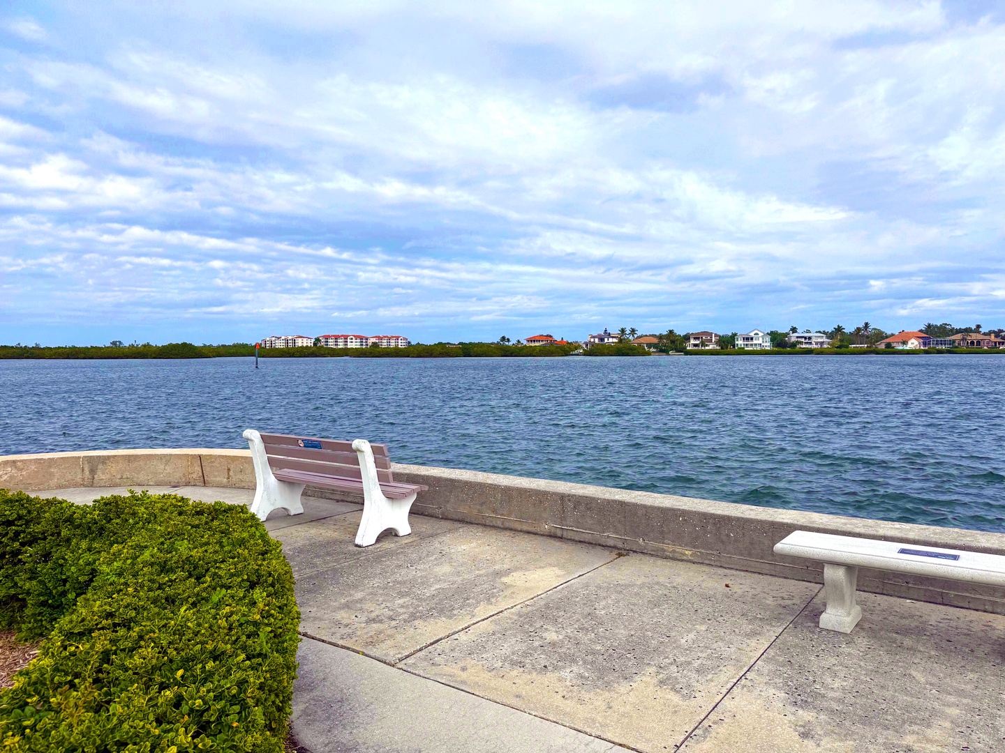 Waterfront seating area with benches overlooking the bay and residential coastline under expansive cloudy skies.