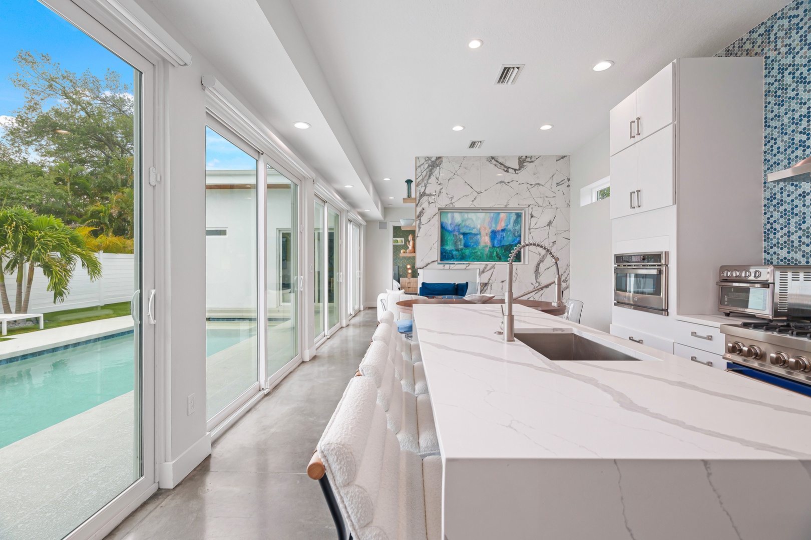 Step into your bright, open kitchen where white marble countertops gleam and floor-to-ceiling windows frame the sparkling pool just beyond the glass.