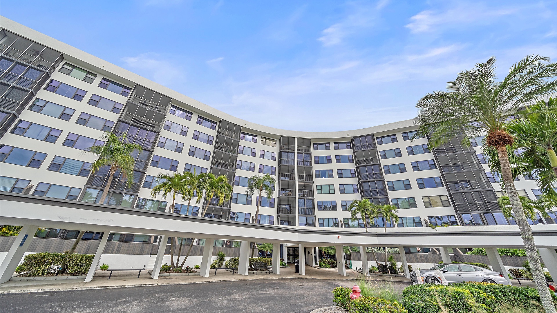 Modern curved hotel building with contemporary architecture, surrounded by tropical palm trees and landscaped grounds in a sunny location.