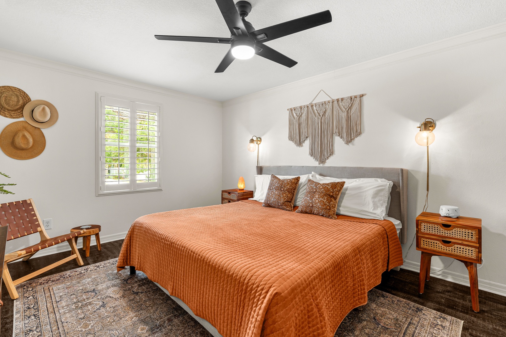 Drift off in your bright, bohemian-inspired bedroom with warm linens, modern ceiling fan, natural light, and thoughtful touches like macramé art and cozy lounge chair.