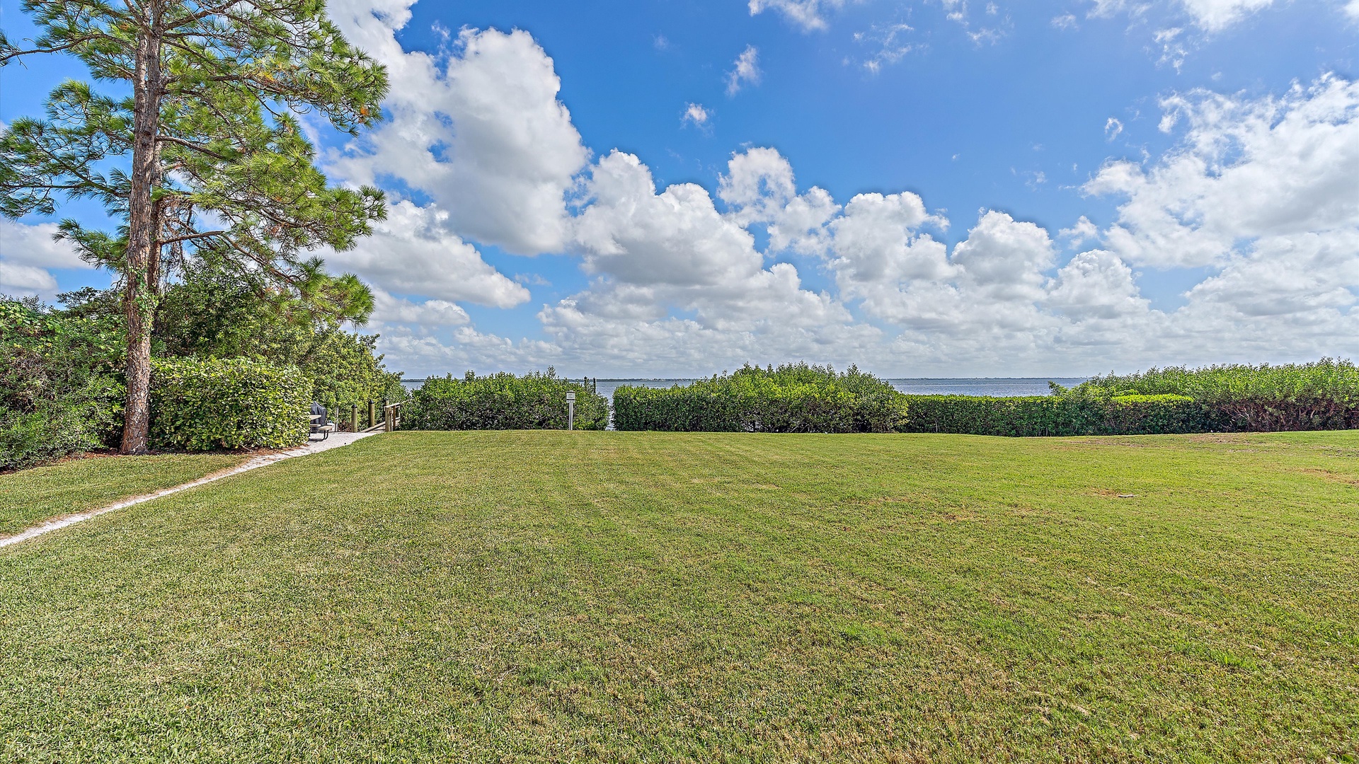 A well-maintained grassy field with a tree on the left, bordered by bushes and shrubs, with a view of the ocean and a partly cloudy sky in the background.