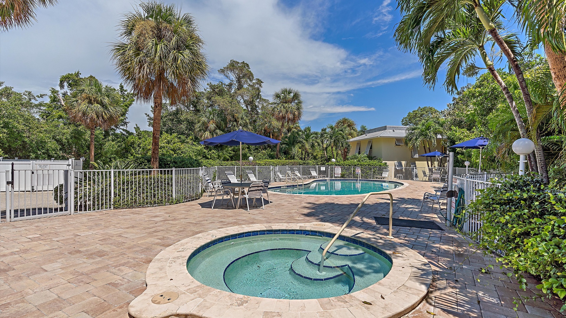 Outdoor pool area with a hot tub in the foreground and a swimming pool in the background. The area features lounge chairs, blue umbrellas, and is surrounded by lush greenery and palm trees.