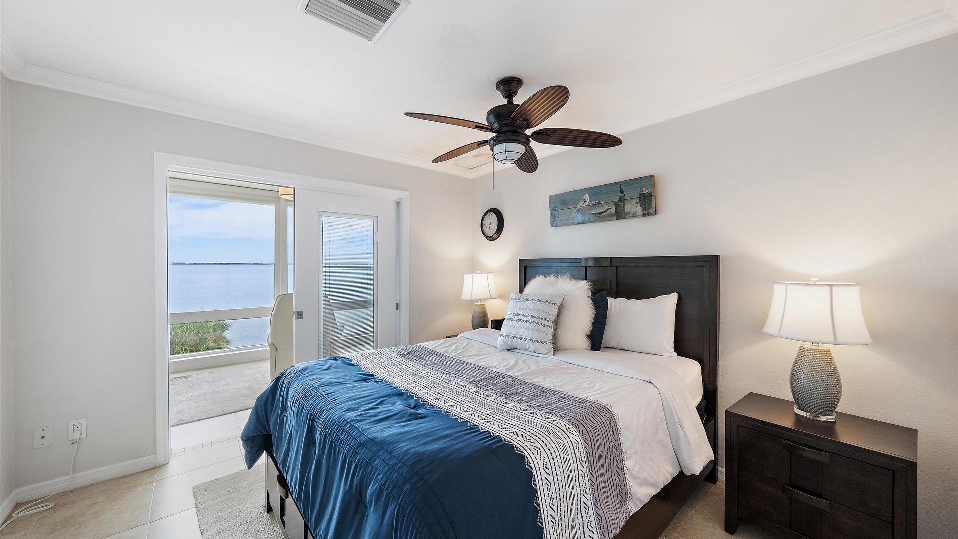 A welcoming queen bedroom with crisp blue-and-white bedding and warm wood furnishings. Access to the enclosed sunroom brings the waterfront atmosphere into this inviting second bedroom.