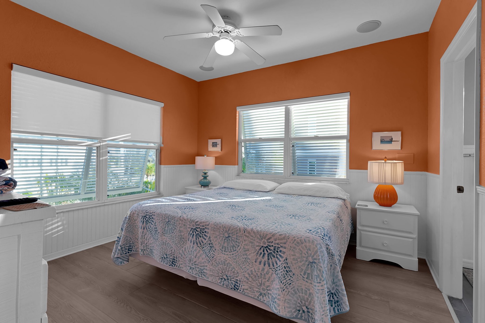 Drift off in your cheerful bedroom featuring warm orange walls, crisp white wainscoting, and breezy windows that invite natural light to dance across the polished floors.