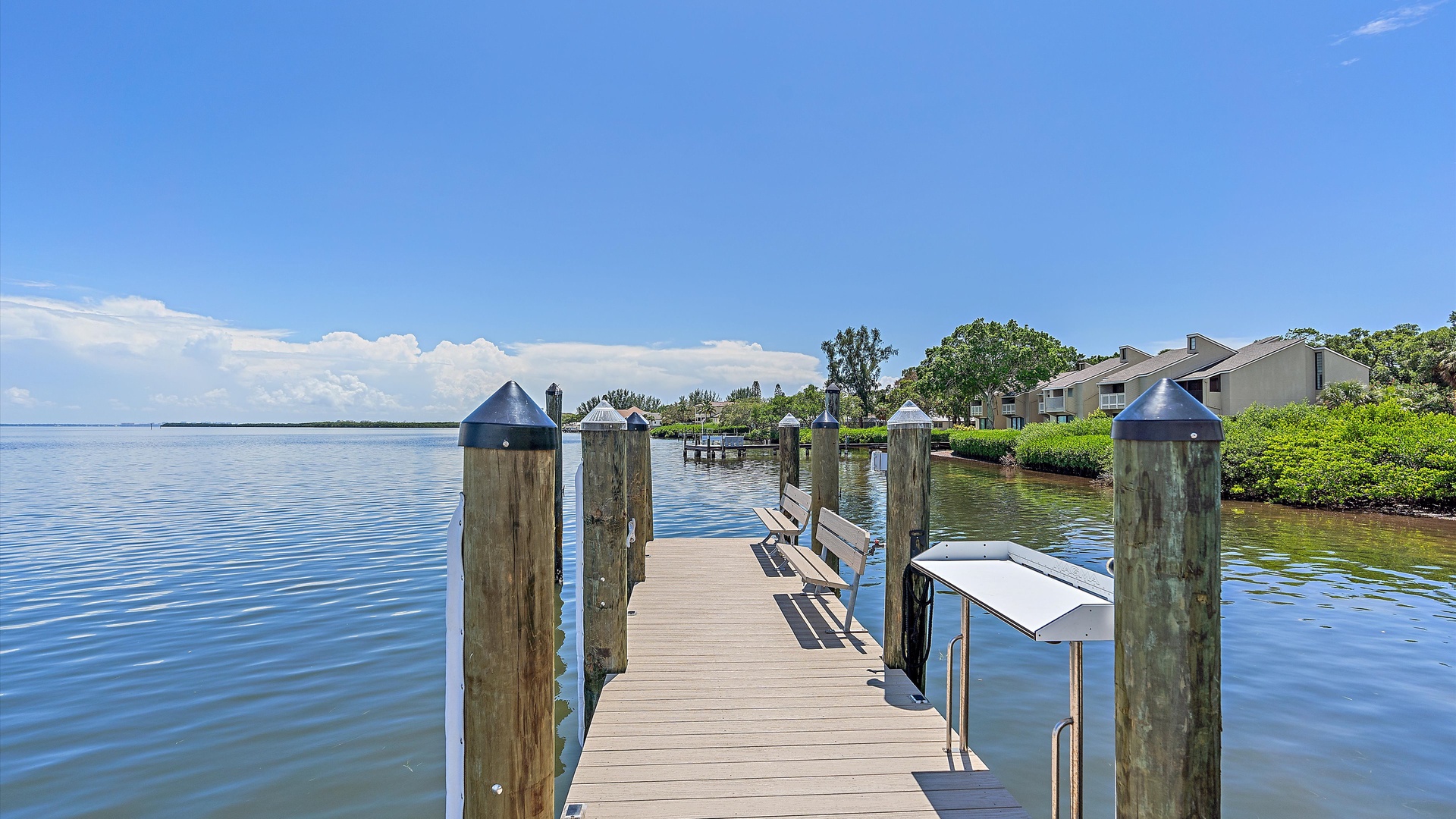 A wooden dock extends into a calm body of water with several benches and a fish cleaning station. Residential houses and trees are visible in the background under a clear, sunny sky.