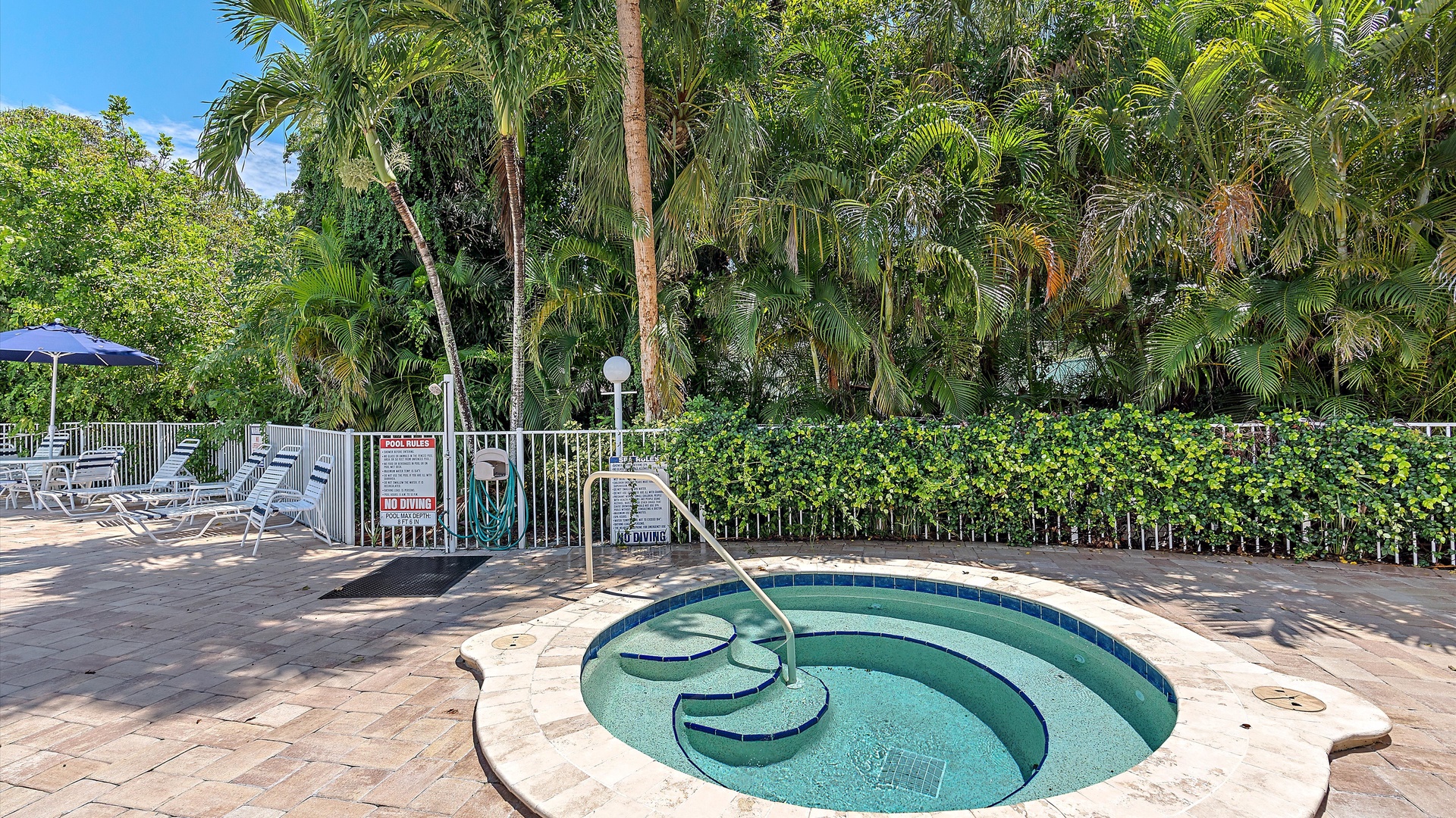 A round hot tub is set in a paved area with lounge chairs and an umbrella nearby, surrounded by a metal fence and lush greenery.