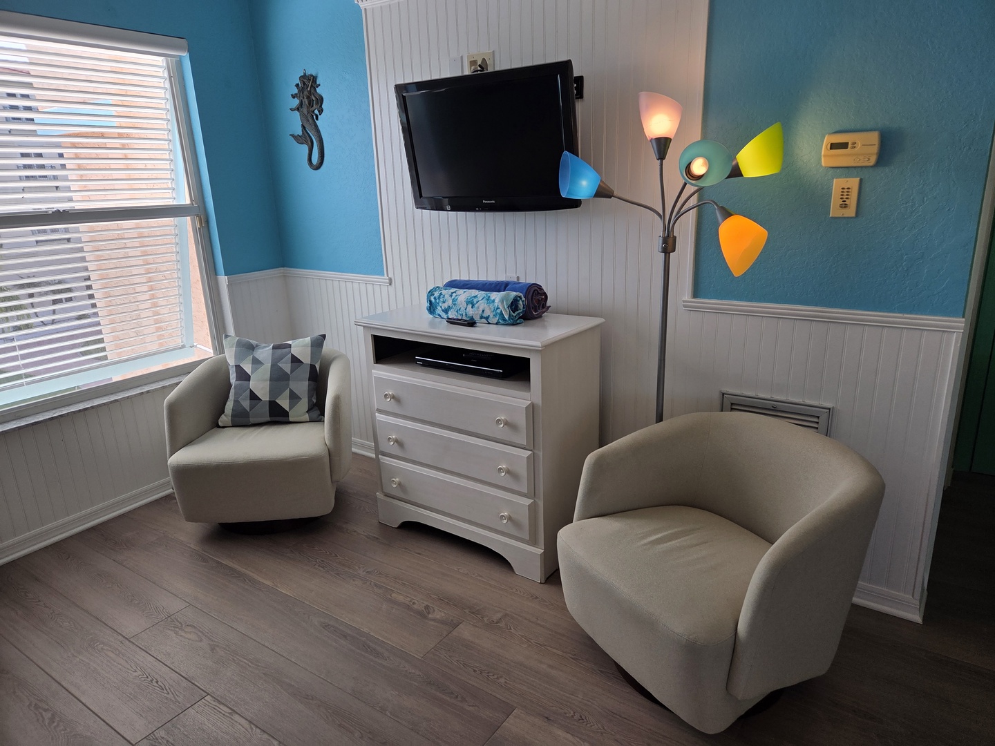 Your bright coastal sitting area features cheerful teal walls and comfy seating perfect for unwinding. The multi-color floor lamp adds a playful touch while the window brings in natural light.