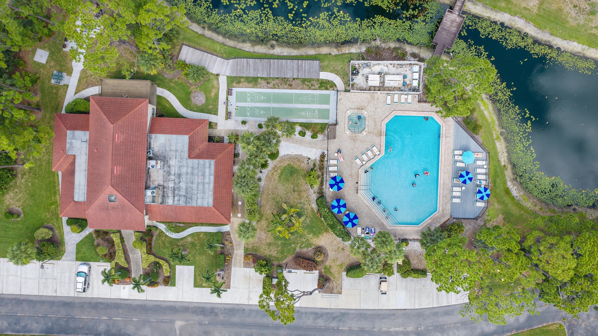 Aerial view of the vacation rental property showcasing the main building with red-tile roof, outdoor swimming pool with lounge chairs, tennis court, natural pond, and lush landscaping throughout the grounds.