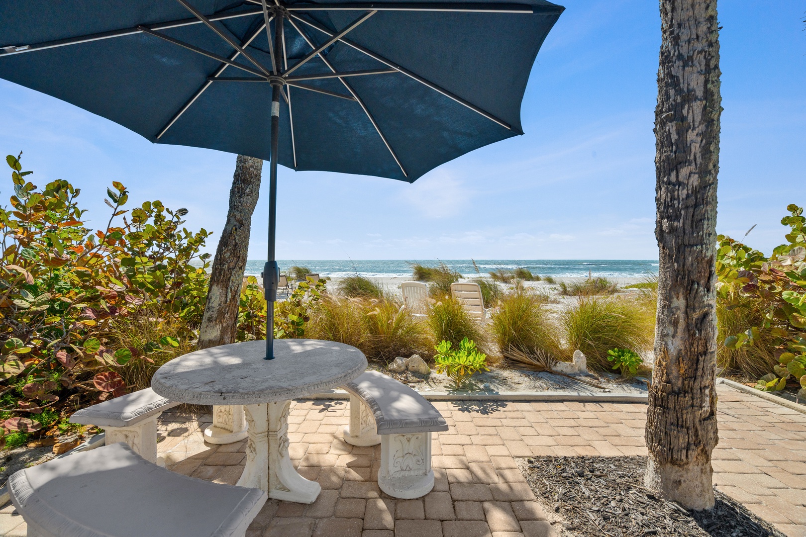 Beachfront patio with stone dining table, umbrella shade, and direct ocean views through tropical landscaping.