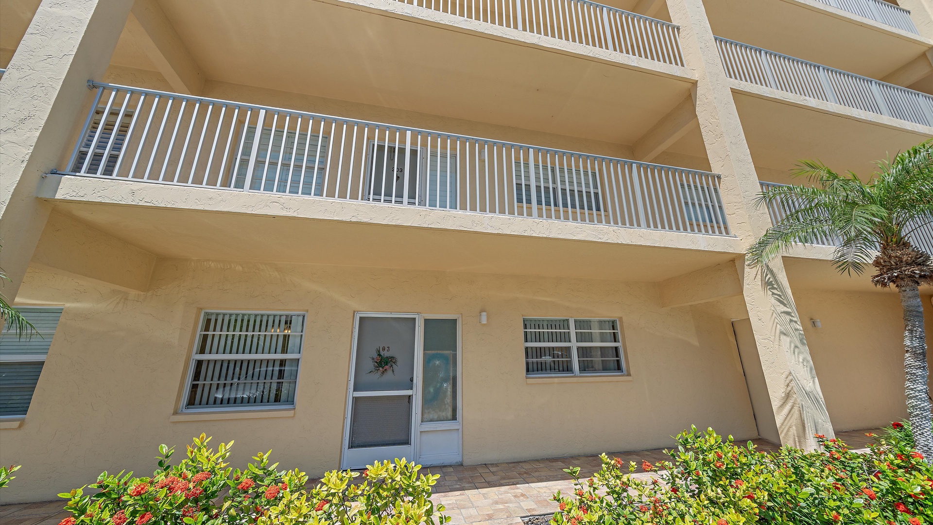 Sunny beachside building with private balconies and tropical landscaping, featuring your ground-floor entrance surrounded by vibrant flowering plants and swaying palms.