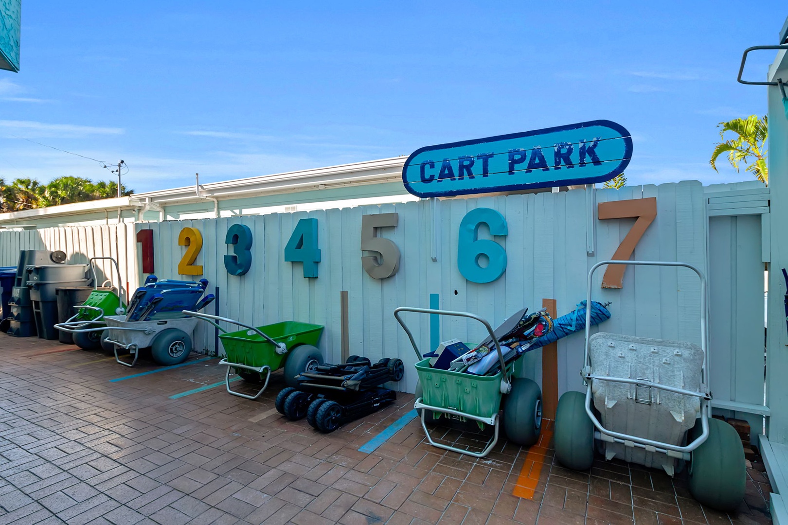 The property's cart park features numbered parking spaces for beach carts and water sports equipment against a vibrant turquoise building, with palm trees and blue sky completing the coastal setting.