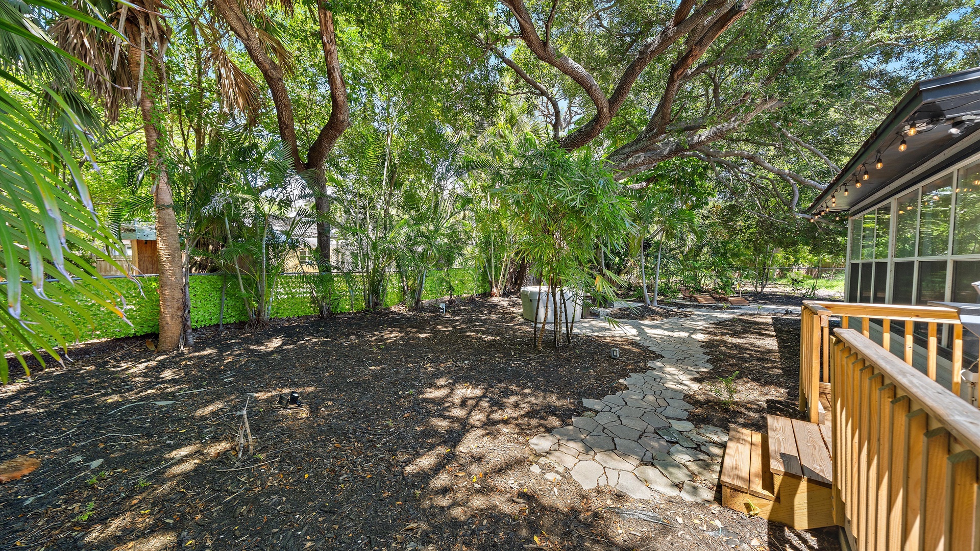 This lush backyard features mature trees creating dappled shade, a natural stone pathway, and a deck extending from the property—your own private nature escape for peaceful mornings or afternoon relaxation.