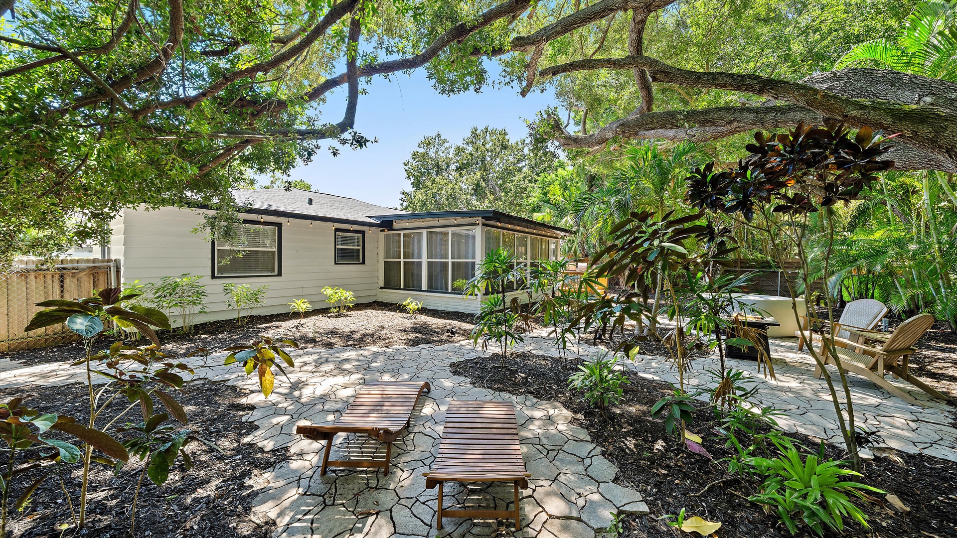 A tranquil exterior view of the property featuring a lush, tropical garden with mature trees, native plantings, and inviting seating areas on a stone patio.