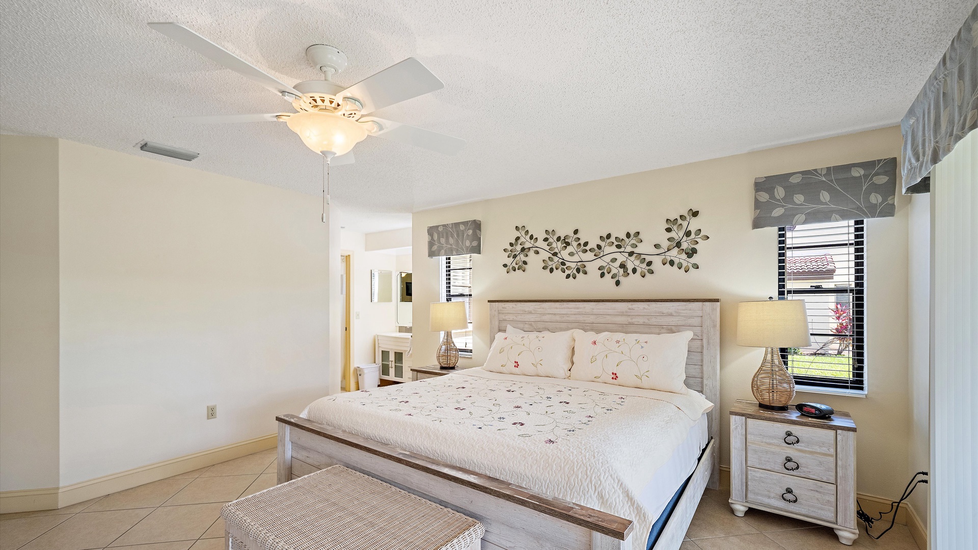 Unwind in your serene retreat where a plush bed, soft lighting, and charming decor invite you to relax. A ceiling fan keeps you cool, and natural light flows through the windows, framing your peaceful stay.