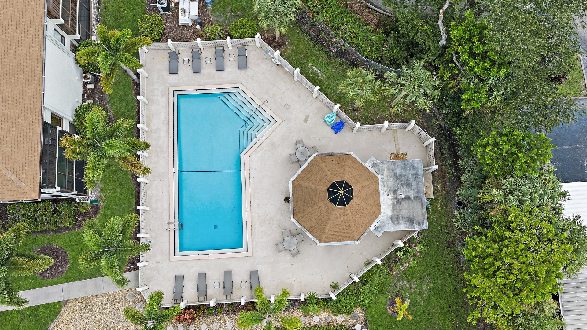 Aerial view of the property's outdoor amenities featuring a swimming pool, sundeck with lounge chairs, and covered seating area surrounded by lush tropical landscaping and garden spaces.