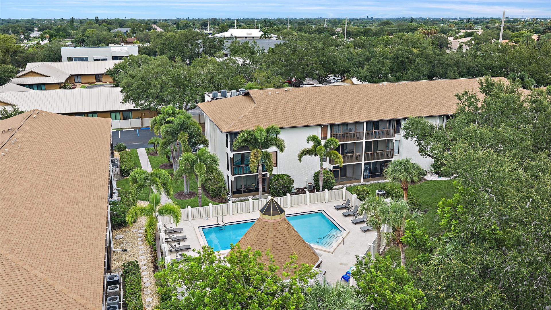 Aerial view of the property showcasing the multi-story vacation rental building, dual swimming pools with surrounding deck, tropical landscaping with palm trees, and lush green neighborhood setting.