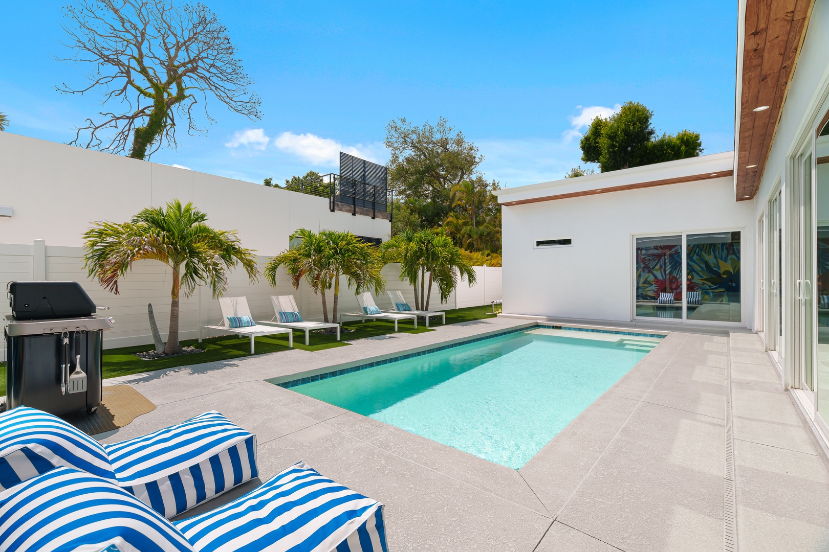 Modern pool area surrounded by tropical palms, sun loungers, and a BBQ grill—perfect for relaxing or entertaining under sunny skies.