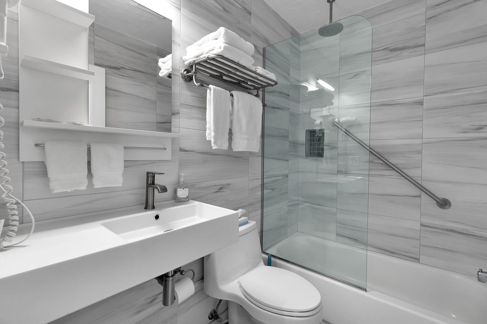 Your modern bathroom features soft wood-look tile, crisp white fixtures, and a luxurious glass-enclosed tub. Fresh towels await you, and the rainfall shower invites relaxation.